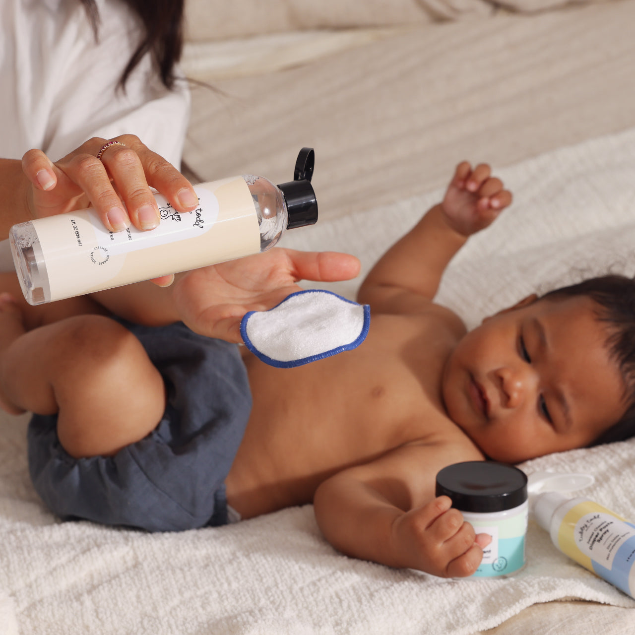 Baby lying on a bed with Mama holding a bottle of Micellar Water, surrounded by Tubby Todd products.